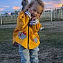 casual_clothing, child, clouds, evening, fence, fun, grass, grass_field, happy, jeans, outdoor, person, playful, pose, sky, smile, sunset, white_shoes, yellow_jacket, young_child