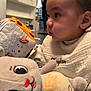 Taylan participe au concours pour gagner de l'argent avec cette photo : baby, child, stuffed_toy, plush, indoor, cozy_clothing, curious, closeup, face, hand, soft_toys, child_playing, person_in_background, home_interior, furniture, shelf, blurred_background, cute, young_child, infant