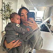 Taylan participe au concours pour gagner de l'argent avec cette photo : woman, baby, mirror, selfie, grey_sweater, baby_clothes, kitchen, potted_plant, modern_interior, reflection, phone, holding, casual_clothing, indoor, portrait, person, home, light, window, floor