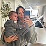 woman, baby, mirror, selfie, grey_sweater, baby_clothes, kitchen, potted_plant, modern_interior, reflection, phone, holding, casual_clothing, indoor, portrait, person, home, light, window, floor
