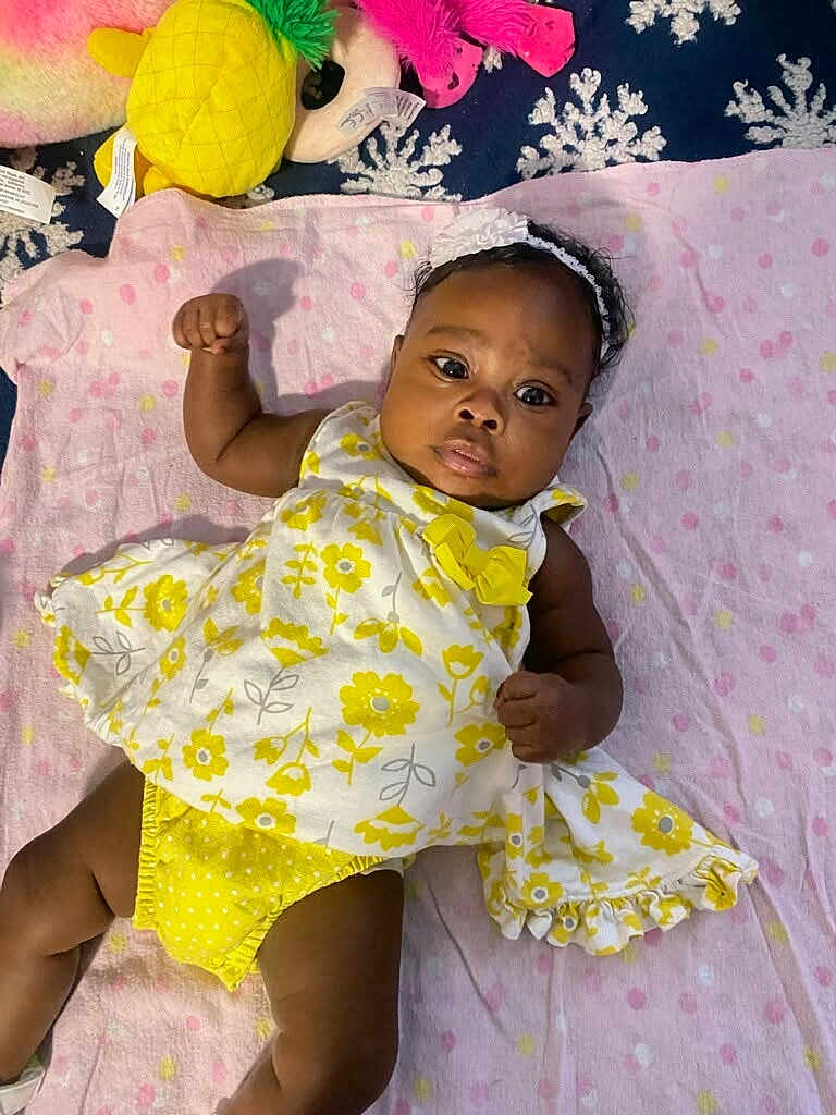 Kalani is registered to the contest to win money with this photo: baby, infant, girl, yellow_dress, headband, blanket, plush_toys, pineapple_toy, pink_flamingo_toy, lying_down, polka_dot, cute, child, indoors, soft_toys, playful, young, portrait, colorful, smiling