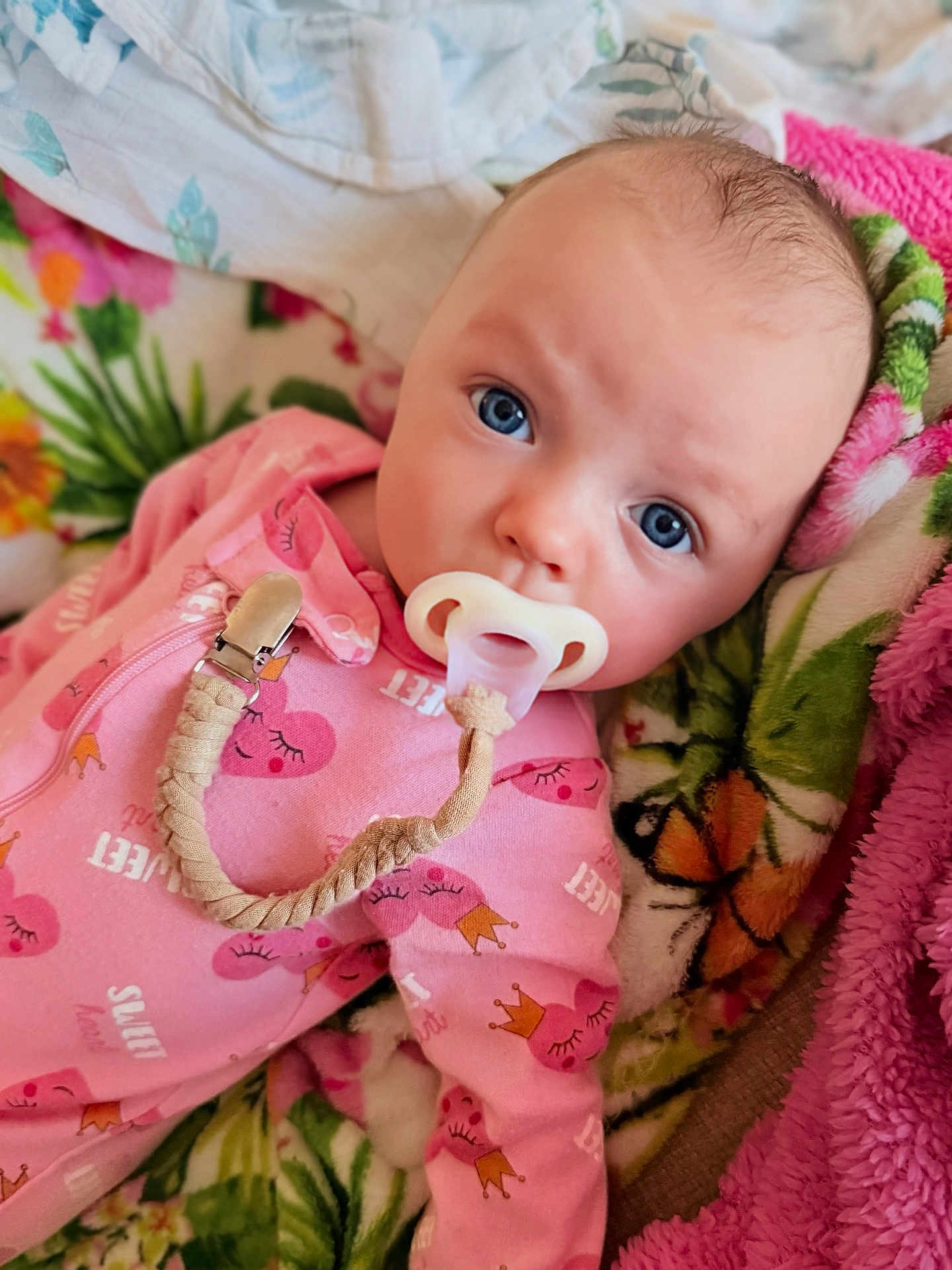 Evelynne joined the competition — help win amazing prizes! baby, infant, pacifier, pacifier_clip, pink_onesie, blue_eyes, blanket, floral_blanket, braided_strap, clip, close_up, portrait, lying_down, cozy, soft_texture, newborn, cute, gaze, bed_bedding, sleepwear