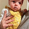 adult_hand, baby, cheeks, child, close_up, clothing, cute, eyes, face, floral, hand, headband, holding, indoors, infant, mouth, portrait, shoulder, skin, yellow_romper