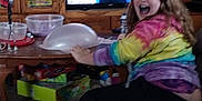 Savannah is registered to the contest to win money with this photo: child, glasses, tie_dye_hoodie, balloon, living_room, carpet, wooden_cabinet, board_games, television, smile, playing, seated, indoor, person, furniture, home, glass, colorful_clothing, braces, happy