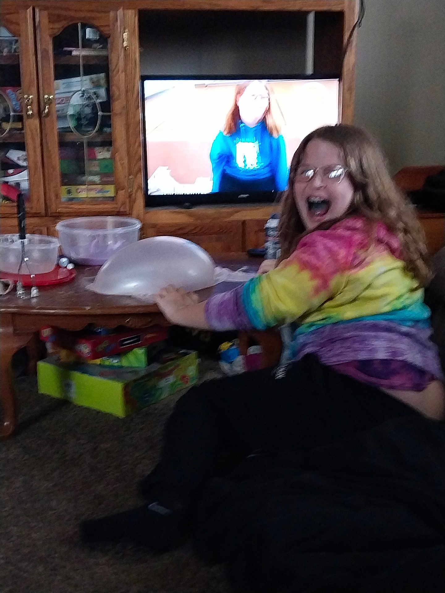 Savannah is registered to the contest to win money with this photo: child, glasses, tie_dye_hoodie, balloon, living_room, carpet, wooden_cabinet, board_games, television, smile, playing, seated, indoor, person, furniture, home, glass, colorful_clothing, braces, happy
