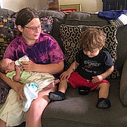 Savannah is registered to the contest to win money with this photo: baby, bottle_feeding, toddler, child, couch, living_room, pillow, blanket, person, sitting, indoor, clothing, glasses, shorts, shirt, footwear, hair, floor, bowl, curious
