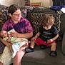baby, blanket, bottle_feeding, bowl, child, clothing, couch, curious, floor, footwear, glasses, hair, indoor, living_room, person, pillow, shirt, shorts, sitting, toddler