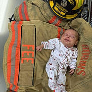 Jace is registered to the contest to win money with this photo: baby, clothing, cute, fabric, firefighter_helmet, firefighter_jacket, helmet_badge, indoor, infant, orange_stripes, person, protective_gear, rescue, resting, safety, sleeping, small, textile, uniform, white_onesie