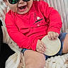 baby, child, sunglasses, hat, red_shirt, blue_shorts, blanket, smiling, happy, infant, cute, clothing, person, hand, sign, indoor, portrait, sitting, cozy, reflection