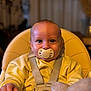 baby, pacifier, high_chair, yellow_clothing, straps, infant, child, face, eyes, hand, indoors, soft_lighting, portrait, cute, cozy, seat, person, young_child, focus, blurred_background
