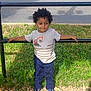 child, toddler, boy, curly_hair, tshirt, pants, sneakers, sidewalk, grass, metal_rail, street, parked_truck, outdoor, daylight, casual_clothing, standing, holding, urban, shadow, concrete