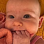 baby, infant, close_up, face, hand, fingers, pink_clothing, yellow_background, skin, chewing, cute, child, portrait, expression, eyes, person, soft_texture, warm_colors, newborn, head