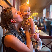 Nora is registered to the contest to win money with this photo: baby, woman, kiss, smile, indoor, people, child, holding, yellow_clothing, denim_vest, happy, face, person, light, blurred_background, chair, table, casual, warm_tone, family