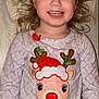 child, smiling, curly_hair, reindeer, festive_clothing, red_nose, santa_hat, long_sleeve_shirt, patterned_shirt, wooden_background, happy, portrait, indoor, cute, holiday_theme, girl, person, face, clothing, red_polka_dot