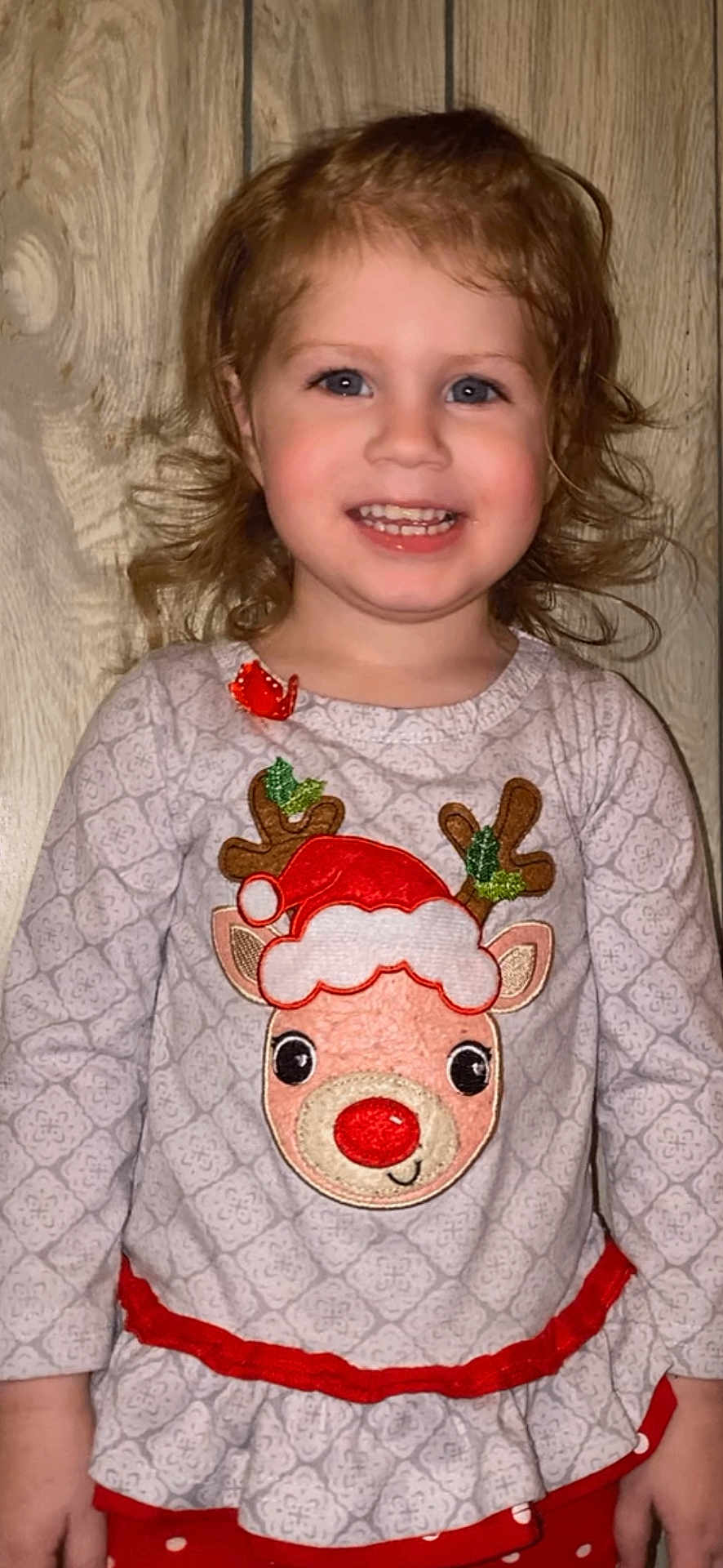 Kinsley is registered to the contest to win money with this photo: child, smiling, curly_hair, reindeer, festive_clothing, red_nose, santa_hat, long_sleeve_shirt, patterned_shirt, wooden_background, happy, portrait, indoor, cute, holiday_theme, girl, person, face, clothing, red_polka_dot