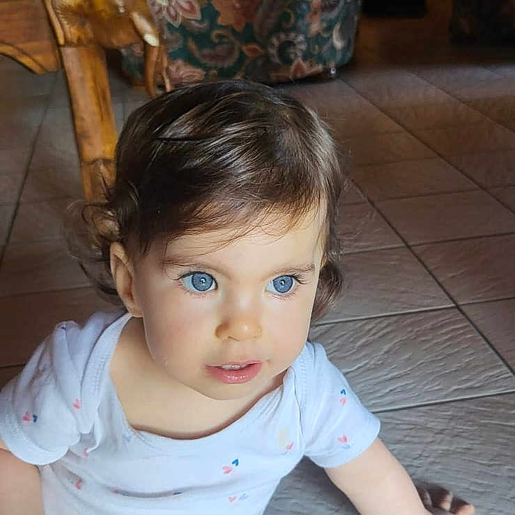 Mia participe au concours pour gagner de l'argent avec cette photo : armchair, blue_eyes, casual_clothing, child, curly_hair, cute, floor_tiles, floral_pattern, happy, home, indoor, natural_light, person, pink_leggings, portrait, sitting, toddler, white_shirt, wooden_chair, young_child