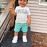 toddler, child, smile, cap, tshirt, shorts, sneakers, porch, railing, house, brick, window, outdoor, happy, person, clothing, footwear, daylight, standing, casual