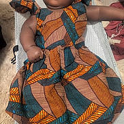 Fatoumata a rejoint le concours — aidez-le/la à gagner de superbes lots ! baby, child, dress, patterned_clothing, sitting, chair, face, indoor, floor, rug, sock, person, infant, cute, portrait, resting, fabric, texture, home, comfort