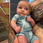 Taytum is registered to the contest to win money with this photo: baby, infant, tattoo, arm, hand, clothing, onesie, pattern, turtle, person, indoor, tile, wall, sign, staircase, curious, holding, skin, adult, portrait