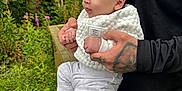 Taytum is registered to the contest to win money with this photo: baby, person, child, tattoo, hand, clothing, white_sock, white_pants, white_jacket, outdoor, wooden_railing, greenery, flowers, forest, nature, portrait, holding, cute, curious, infant