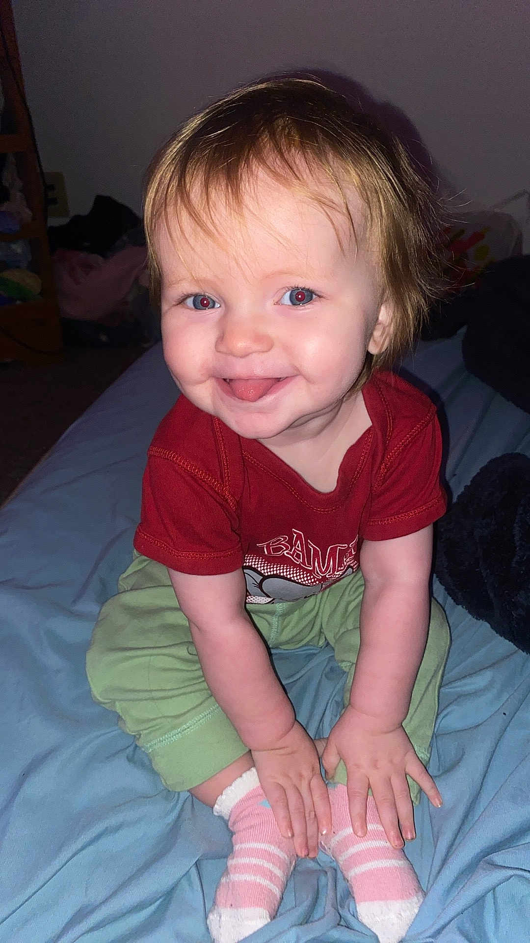 Delilah joined the competition — help win amazing prizes! baby, child, smiling, tongue_out, sitting, clothing, red_shirt, green_pants, pink_socks, bed, blanket, indoor, human, cute, happy, portrait, toddler, hands, face, hair