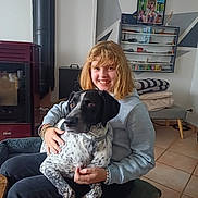 Otto participe au concours pour gagner de l'argent avec cette photo : girl, person, dog, pet, animal, smiling, portrait, indoors, sitting, cuddling, black_and_white, speckled, long_hair, sweatshirt, bracelet, chair, fireplace, shelves, framed_photos, home