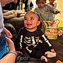 baby, child, smiling, skeleton_outfit, socks, person, indoor, toy, furniture, adult, phone, happy, playing, living_room, couch, colorful, curly_hair, casual_clothing, interaction, family