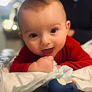 Milo a rejoint le concours — aidez-le/la à gagner de superbes lots ! baby, background_blur, child, close_up, cute, expression, face, focus, hand, happy, indoor, infant, lying_down, person, playful, red_clothing, skin, smiling, soft_lighting, toy
