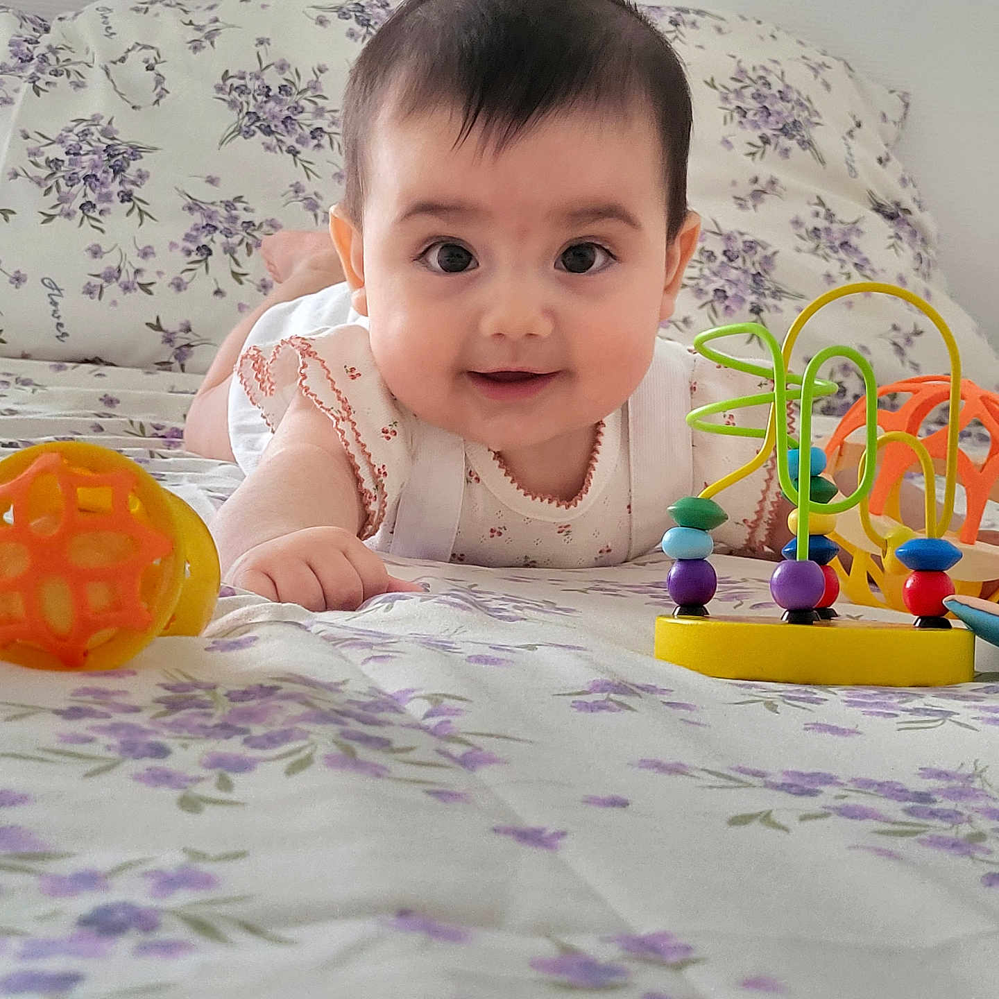 Martina a rejoint le concours — aidez-le/la à gagner de superbes lots ! baby, bed, bright, cheerful, child, closeup, clothing, colorful, cute, face, floral_bedspread, happy, indoors, infant, lying_down, person, playtime, smiling, toy, young