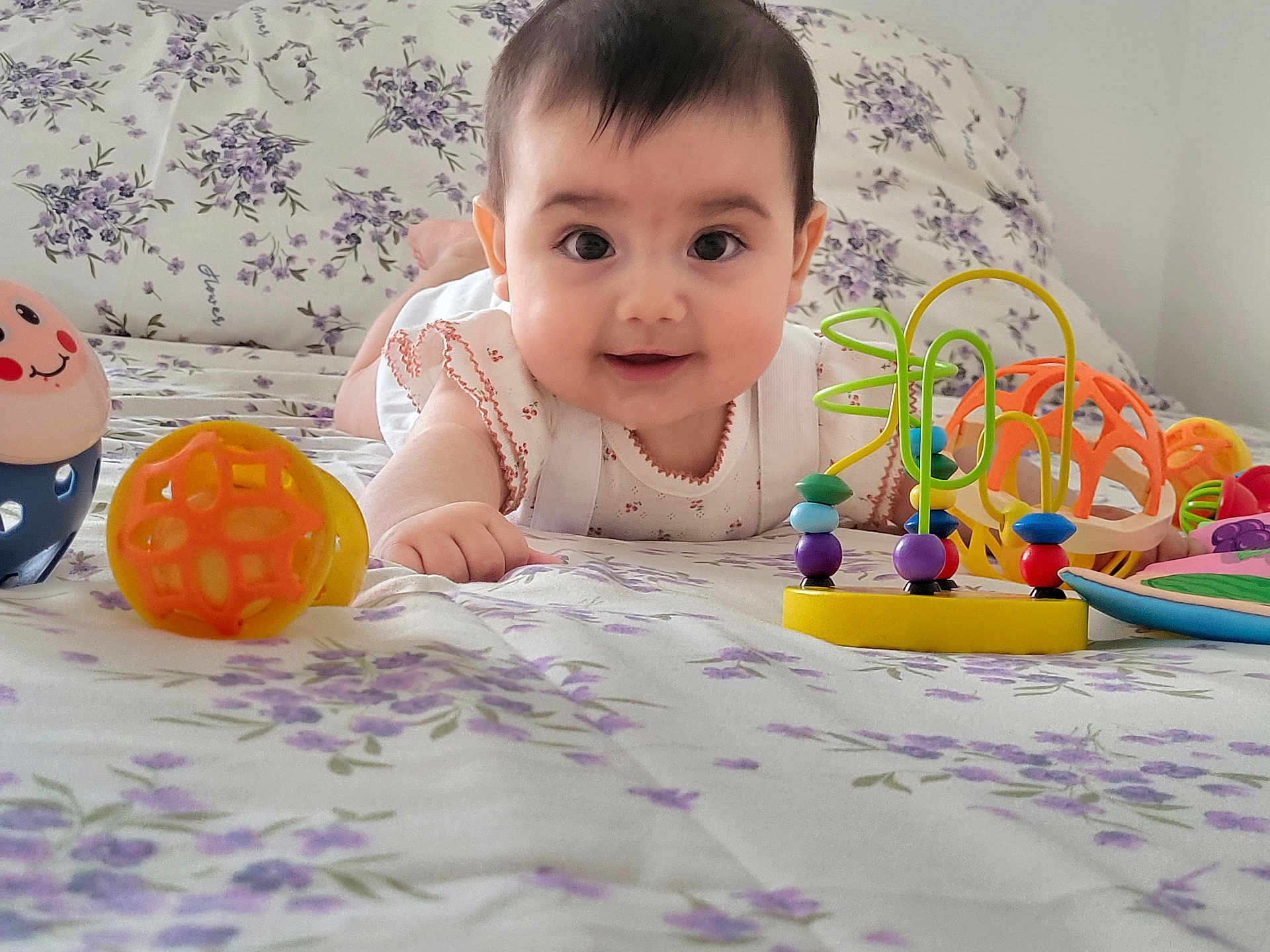 Martina a rejoint le concours — aidez-le/la à gagner de superbes lots ! baby, bed, bright, cheerful, child, closeup, clothing, colorful, cute, face, floral_bedspread, happy, indoors, infant, lying_down, person, playtime, smiling, toy, young