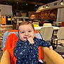 Kleos a rejoint le concours — aidez-le/la à gagner de superbes lots ! baby, child, high_chair, smiling, indoor, cafe, wooden_chair, orange_jacket, clothing, happy, person, table, modern_interior, lighting, face, finger_in_mouth, seatbelt, casual_wear, cute, young_child