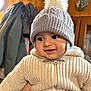 baby, child, knit_hat, pom_pom, sweater, knitwear, indoor, wooden_wall, coat, hand, smile, cute, warm_clothing, face, person, winter, apparel, headwear, happy, portrait