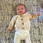 Eden a rejoint le concours — aidez-le/la à gagner de superbes lots ! baby, infant, smiling, lying_down, beige_clothing, white_pants, patterned_bedspread, cute, happy, child, portrait, indoor, cozy, buttoned_sweater, small_hands, head, face, young, resting, toddler