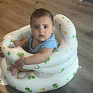 Abdulmajid is registered to the contest to win money with this photo: baby, blue_clothing, child, curious, cute, expression, floor, frog_pattern, home, indoor, infant, inflatable_seat, looking, moon_pattern, person, seat, sitting, toy_pattern, wooden_floor, young_child