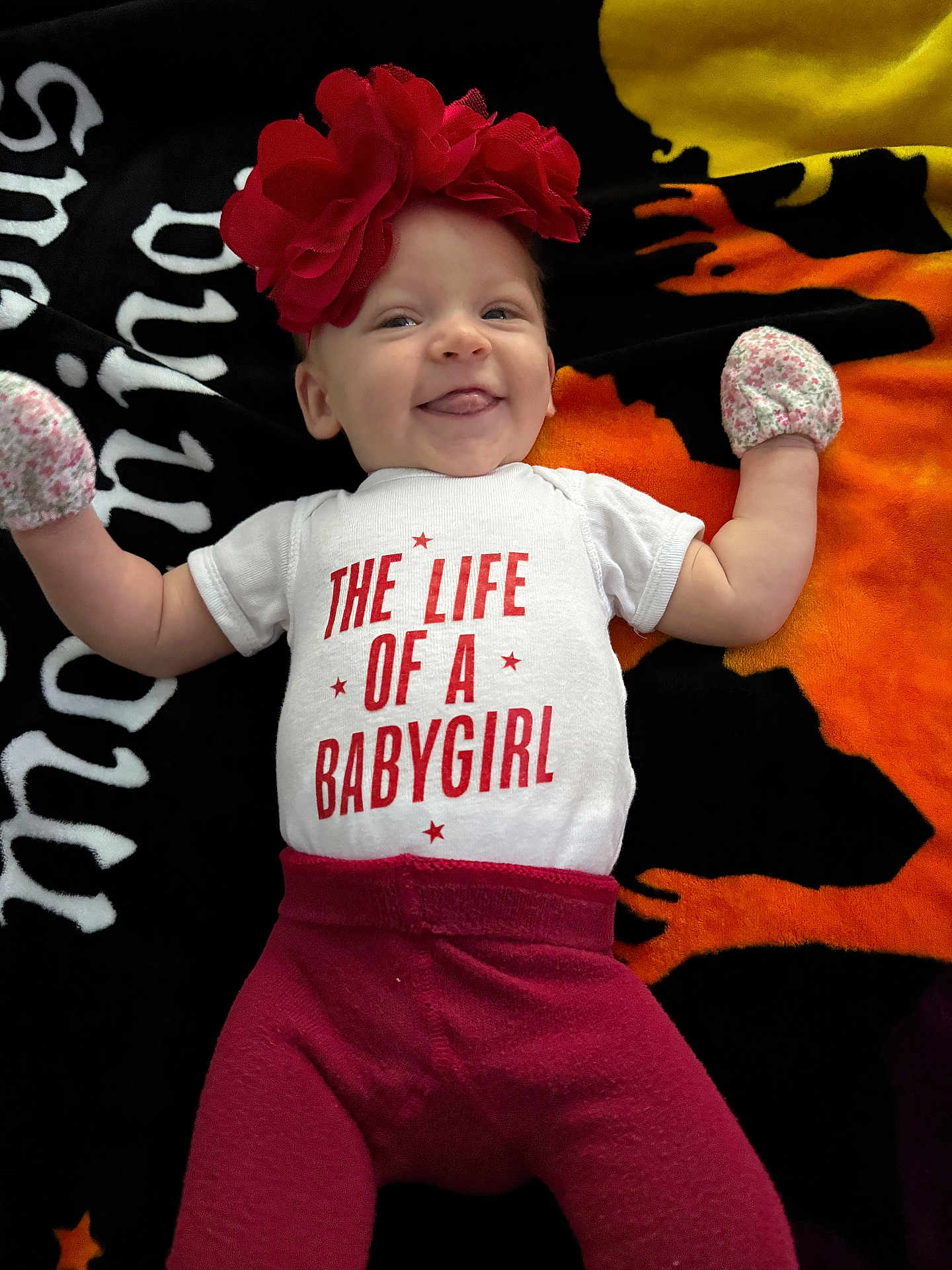 Brooklynn is registered to the contest to win money with this photo: baby, infant, smiling, red_flower_headband, mittens, white_onesie, red_pants, tongue_out, lying_down, colorful_blanket, halloween_theme, orange, black, yellow, cute, child, portrait, happy, playful, indoors