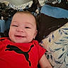 baby, smile, red_shirt, puma_logo, blanket, bed, blue, brown, face, child, happy, infant, cute, lying_down, indoors, person, young, soft, cozy, portrait