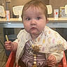 baby, child, high_chair, bib, cardigan, food, french_fries, table, indoor, restaurant, face, curious, seated, person, clothing, expression, wooden_table, cute, infant, dining