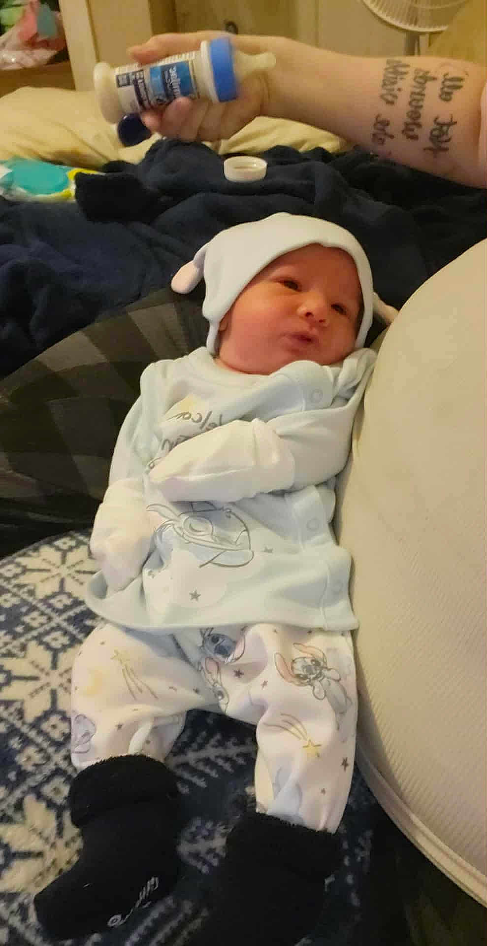Greyson is registered to the contest to win money with this photo: baby, newborn, pajamas, hat, bottle, hand, blanket, cushion, indoor, person, infant, footwear, carpet, pattern, clothing, sleepwear, cute, relaxing, resting, home