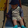 child, toddler, slide, playground, hat, socks, cardigan, overalls, metal_slide, gravel, outdoor, daylight, yellow_playground_equipment, person, baby, seat, sunlight, shadow, casual_clothing, nature