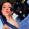 woman, person, puppy, dog, black_dog, pet, car, selfie, portrait, kissy_face, tongue_out, fluffy, bow, purple_bow, blue_outfit, seatbelt, vehicle, indoor, daylight, cuddling