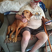 Duke Thomas is registered to the contest to win money with this photo: man, dog, couch, cap, glasses, phone, cane, blanket, poster, wooden_cane, shorts, tshirt, footwear, indoor, pet, seated, relaxed, living_room, beard, smiling