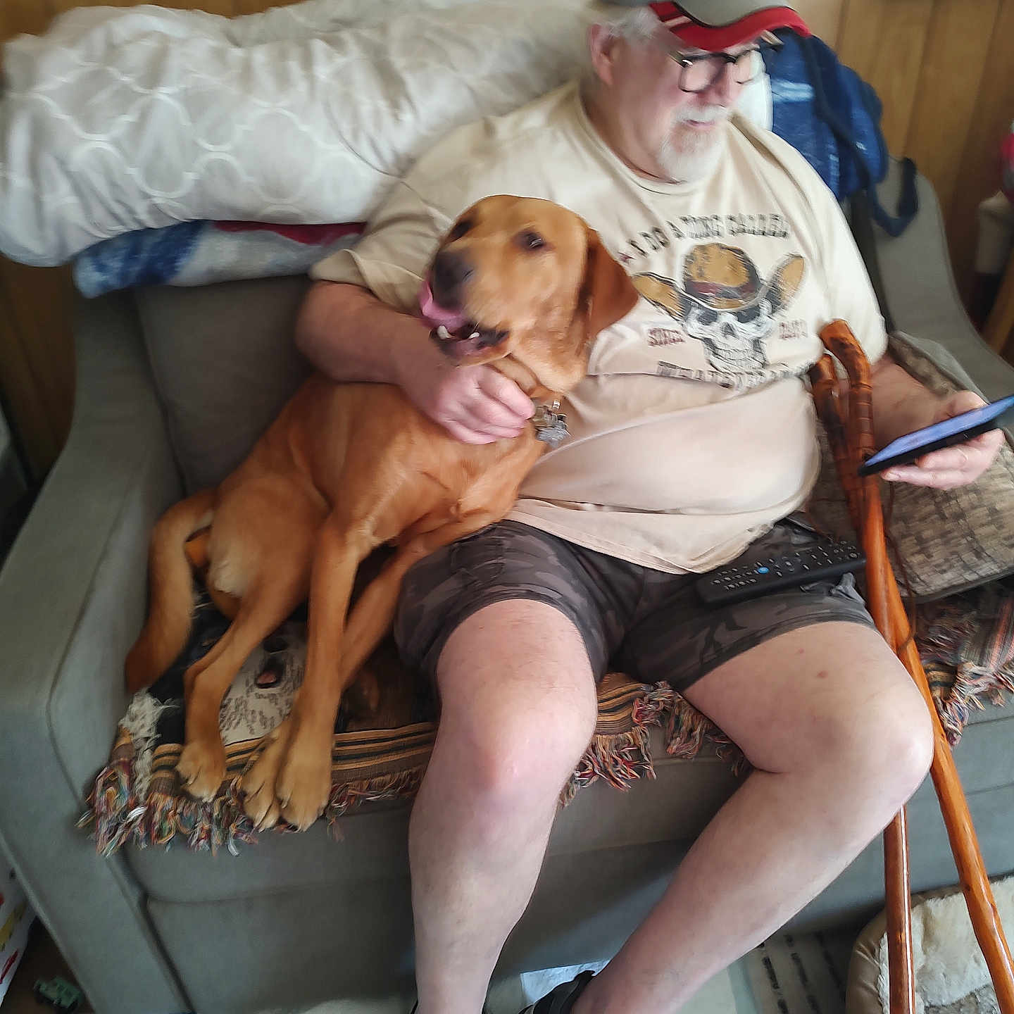 Duke Thomas is registered to the contest to win money with this photo: man, dog, couch, cap, glasses, phone, cane, blanket, poster, wooden_cane, shorts, tshirt, footwear, indoor, pet, seated, relaxed, living_room, beard, smiling