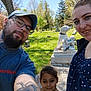 blue_sky, casual_clothing, cemetery, child, dog_statue, family, glasses, grass, headshot, man, nature, outdoor, portrait, selfie, smile, statue, sunny, tattoos, tree, woman