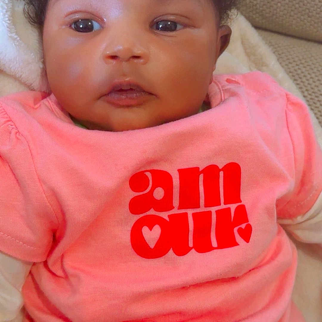 Oumou Koultoumy a rejoint le concours — aidez-le/la à gagner de superbes lots ! adorable, baby, blanket, cheeks, closeup, clothing, couch, curly_hair, eyes, face, gaze, indoor, infant, open_mouth, photography, pink_shirt, portrait, reclining, skin, young_child