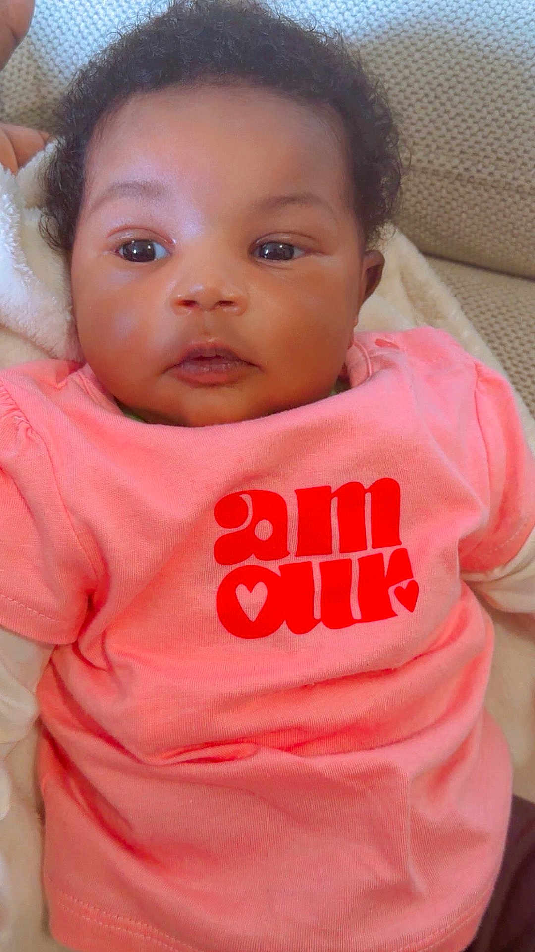 Oumou Koultoumy a rejoint le concours — aidez-le/la à gagner de superbes lots ! baby, infant, face, eyes, curly_hair, pink_shirt, clothing, blanket, couch, closeup, portrait, cheeks, skin, open_mouth, gaze, indoor, reclining, adorable, young_child, photography