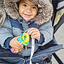 baby, bundle, child, coat, cute, face, fur_trim, happy, holding, hood, infant, jacket, outdoor, person, playful, smiling, stroller, toy, warm, winter_clothing