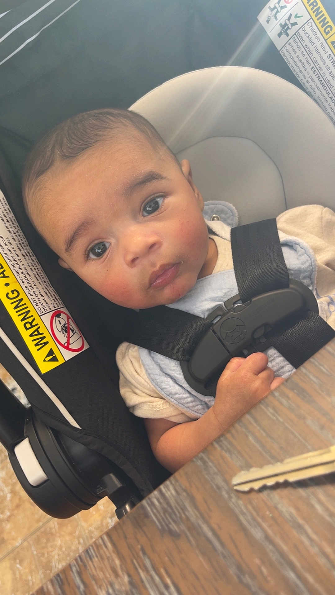 Tommy James is registered to the contest to win money with this photo: baby, child, car_seat, seatbelt, key, wooden_table, indoor, curious, infant, safety, light_glare, face, person, young_child, clothing, hand, closeup, seat, warning_label, expression