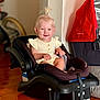 baby_clothing, black_car_seat, blond_hair, car_seat, child, cute, face, floor, happy, home_interior, indoor, infant, person, pony_tail, red_cloth, sitting, smile, stroller, toddler, wooden_floor