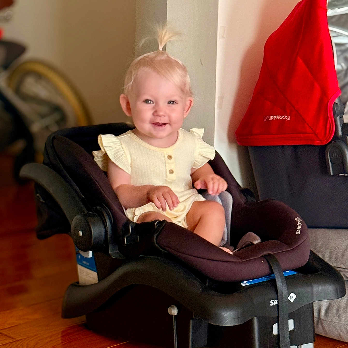 Oliviia is registered to the contest to win money with this photo: baby_clothing, black_car_seat, blond_hair, car_seat, child, cute, face, floor, happy, home_interior, indoor, infant, person, pony_tail, red_cloth, sitting, smile, stroller, toddler, wooden_floor