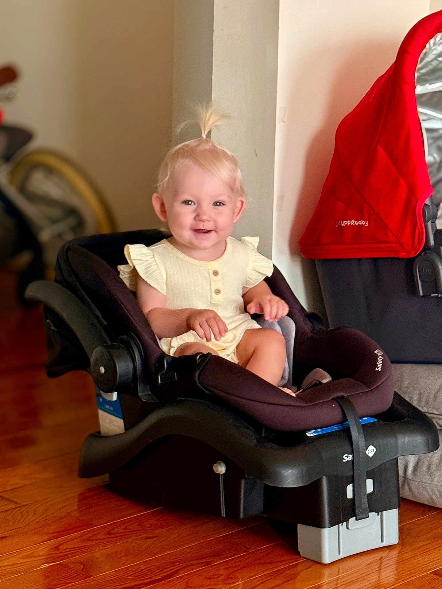 Oliviia is registered to the contest to win money with this photo: baby_clothing, black_car_seat, blond_hair, car_seat, child, cute, face, floor, happy, home_interior, indoor, infant, person, pony_tail, red_cloth, sitting, smile, stroller, toddler, wooden_floor